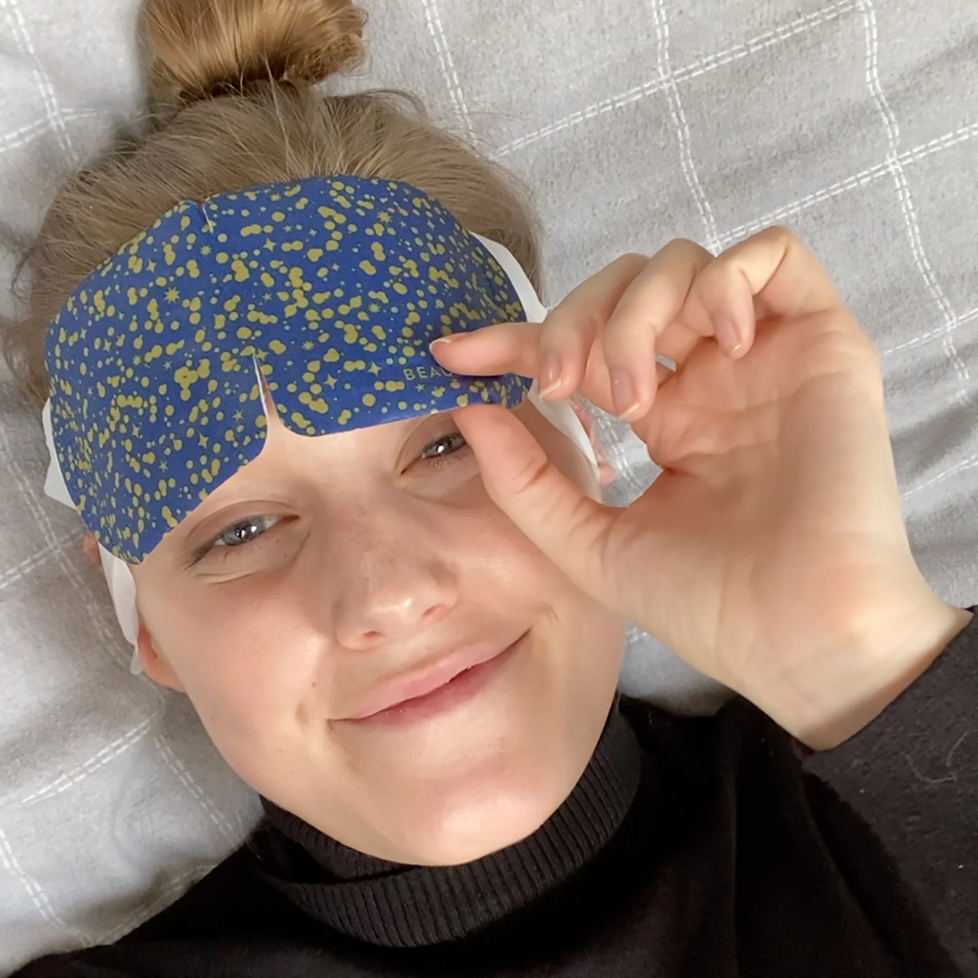 Smiling woman relaxing wearing navy BeautyPro eye mask with gold print while lying down
